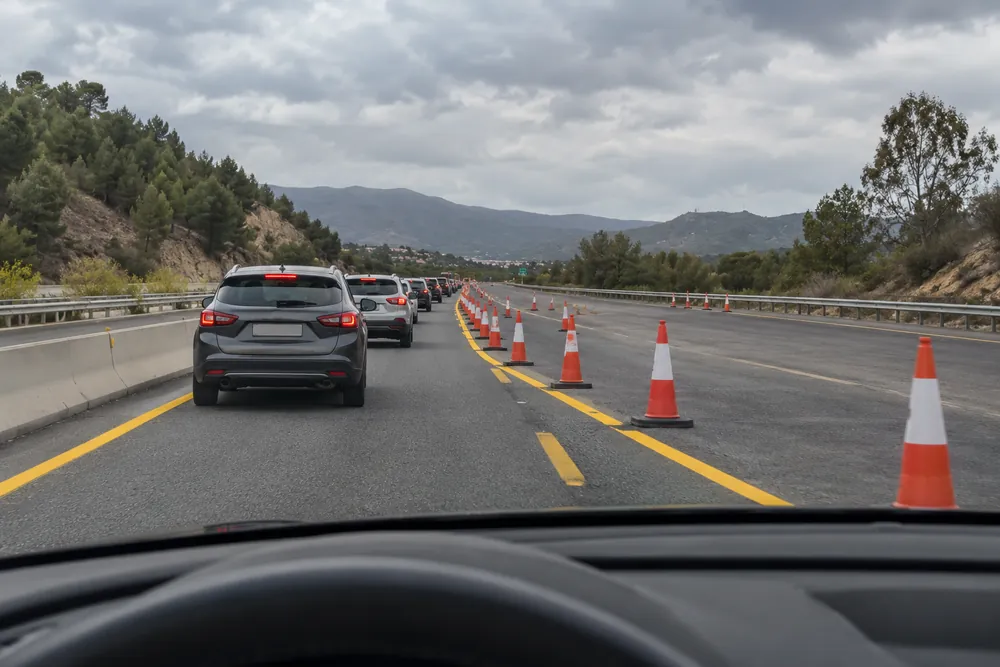Dos carriles que se convierten en uno por obras, con conos y tráfico lento en ambos.