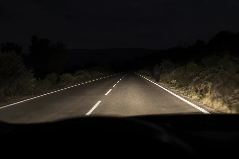 Peatón vestido de oscuro caminando por el borde de una carretera sin iluminar de noche.